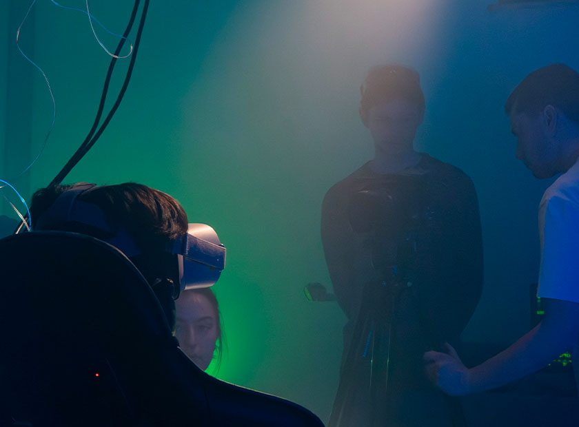 Vue d’ensemble avec trois étudiants : l’un derrière la caméra et l’autre juste à côté, en train de tenir le trépied. Une personne est assise sur un siège, portant un casque de réalité virtuelle sur les yeux.