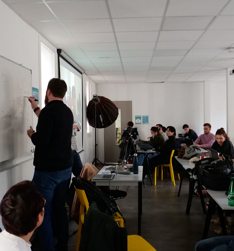 Vue d’ensemble d’une salle de l’ESD avec l’intervenant qui écrit au tableau et les étudiants assis sur leurs chaises.