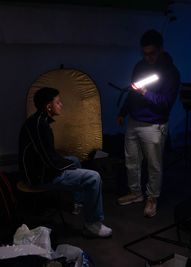 Vue d’une pièce sombre avec deux étudiants : l’un assis sur une chaise, l’autre debout tenant une lampe.