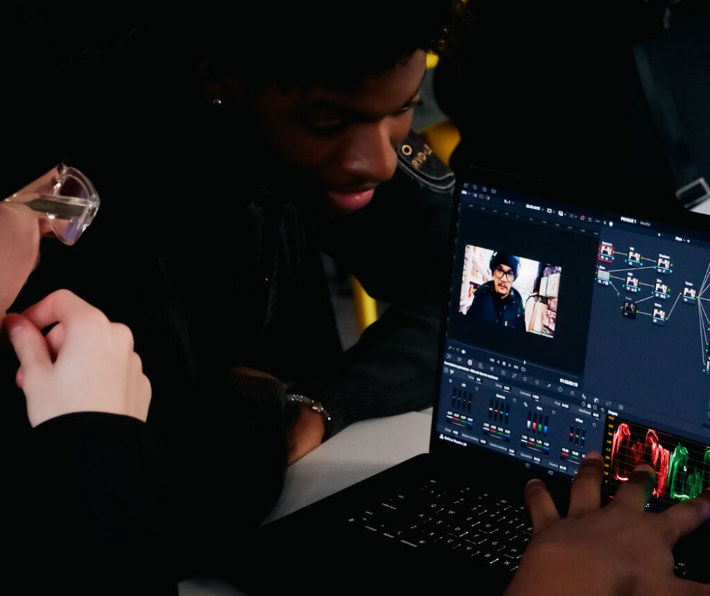 Vue en focus sur un ordinateur affichant un logiciel de montage, avec deux étudiants : l’un face à l’ordinateur, l’autre à côté.