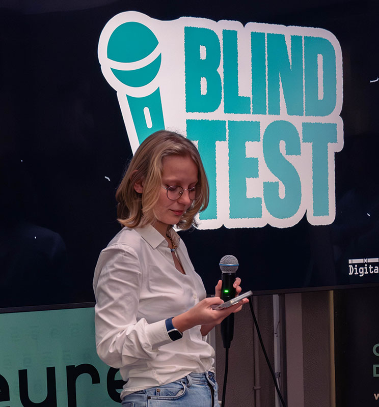 Vue sur une étudiante qui tient un micro, avec derrière elle un écran affichant « Blind Test ».